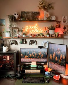 Cozy desk setup with dual monitors. A fall forest as the screen background. Two shelves above the desk with a neon sign that says Good Vibes and cozy gaming decor. Cluttered Gamer Room, Gaming Setup Organization, Maximalist Gaming Room, Cosy Games Room, Eclectic Gaming Room, Cozy Gamer Office, Boho Game Room Ideas, Vintage Gaming Setup Aesthetic, Fall Desk Setup