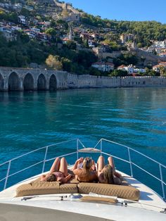 two women lounging on the back of a boat in front of a town and lake