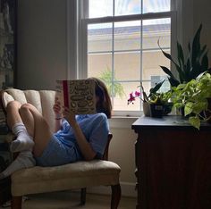 a woman sitting on a chair reading a book in front of a window with potted plants