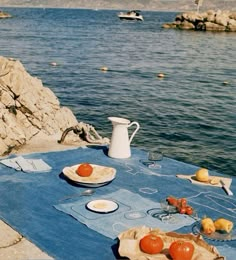 an outdoor table with plates and fruit on it near the water in front of some rocks