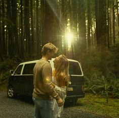 a man and woman standing in front of a van on a forest road with the sun shining through the trees