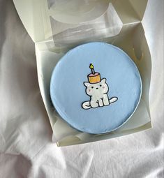 a birthday cake in a box with a cat on it and a candle sticking out of the top