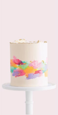 a white cake with multicolored icing on a stand against a pink background