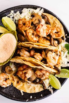 three tacos with shrimp, mushrooms and rice on a black plate next to lime wedges