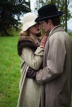 a man and woman standing next to each other in the grass with trees behind them