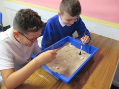 two young boys sitting at a table making sand in a blue tray with spoons