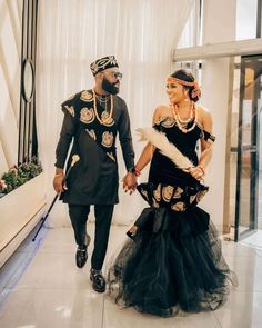 a man and woman dressed in african attire walking down a hall together, holding hands