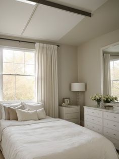 a white bed sitting under a window next to a dresser with drawers and lamps on top of it