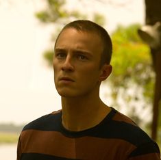 a young man standing in front of a tree looking at the camera while wearing a striped shirt