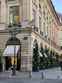 people are walking in front of a large building with christmas trees on the side walk