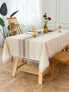 a table with a white table cloth and wooden chairs in front of a plant on the floor