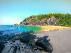 the beach is surrounded by large rocks and green trees in the distance, with clear blue water