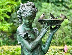 a statue holding a tray with two birds on it in front of some bushes and trees