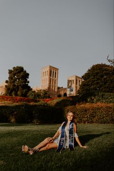 a woman sitting on the grass in front of some bushes and trees with buildings behind her