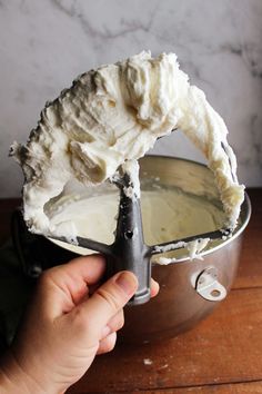 a hand holding a metal bowl filled with white frosting on top of a wooden table