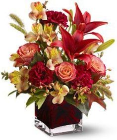 a red vase filled with lots of flowers on top of a white surface and surrounded by greenery