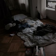 an unmade bed with clothes on the floor next to a window in a dark room