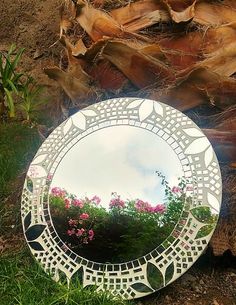 a round mirror sitting on the ground next to some grass and flowers in front of it