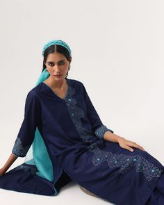 a woman sitting on the ground wearing a blue dress and head scarf with an embroidered design