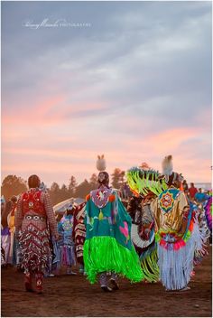 two people dressed in colorful costumes walking down a dirt road at sunset with other people standing behind them