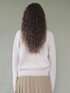 a woman with long brown hair wearing a white sweater and tan pleated skirt standing in front of a gray wall