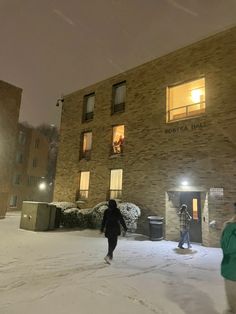 two people are walking in the snow near an apartment building at night with lights on