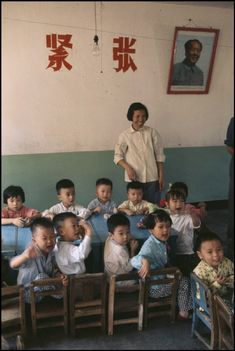 a woman standing in front of a group of children sitting at desks and chairs