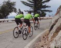 three men in green shirts are riding bikes down the road with large rocks behind them