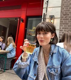 a woman holding up a glass of beer in front of a red building with people sitting at tables