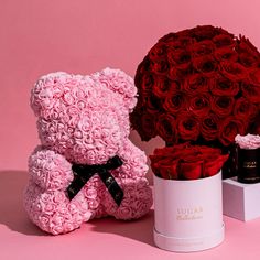 a pink teddy bear next to a large bouquet of red roses and a white box