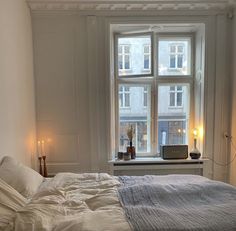 an unmade bed in front of a window with candles on the windowsills