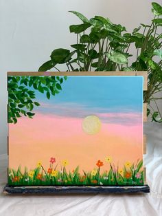 a painting of flowers in the grass with a sunset behind it and a potted plant next to it