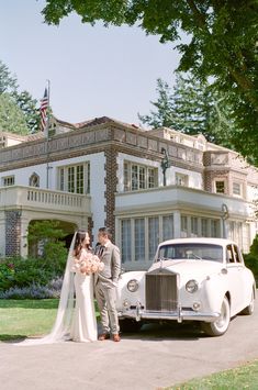 a bride and groom standing in front of a classic car outside a large white house