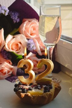 a cupcake with two candles on it sitting next to some flowers and a glass of wine