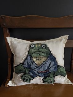 a pillow with a frog on it sitting on top of a wooden chair in front of a black wall