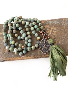 "African Turquoise Mala - 108 Beads - Hand Knotted - Buddha Pendant - 8mm Length Approx. 42\" - 6mm Length Approx. 32\" African Turquoise metaphysical properties are believed to open the mind to new ideas and develop a positive outlook on life. This Mala is made with 108 beads hand knotted between each bead and accented with copper metal spacer beads. This unique Mala that has a removable tassel and seated Buddha pendant. Small Tiger Eye wood beads are added in near the pendant for a earthy desi Green Spiritual Beads, 108 Count, Green Mala With 8mm Beads For Healing, Green Hand-strung Spiritual Mala, Green Spiritual Mala With 108 Beads, Adjustable Green Beaded Necklaces For Meditation, Green Spiritual Beads For Healing, Green Round Meditation Beads, Green Gemstone Beads Mala For Healing, Green 8mm Bohemian Beads