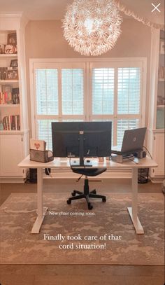 a desk with two computers on it in front of a window and a chandelier