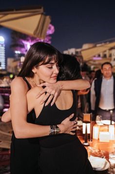 two women hugging each other in front of candles and people at an outdoor dining area