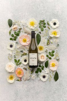 a bottle of wine surrounded by flowers on a white surface with green leaves and petals