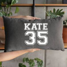 a person holding a pillow with the number 35 on it in front of some plants