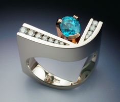 This 14k white and rose gold woman's ring features a beautiful oval Blue Zircon and channel set Diamonds. The design is bold and geometric, with the vibrant Zircon set in rose gold. The Zircon weighs 1.78 carats, and the total diamond weight is .27 carats. The ring weighs 17.6 grams and is a size 6.75. It cannot be sized, but can be custom made in any size. The price of this ring is $4650.00 Modern Rose Gold Jewelry With Tension Setting, Modern Channel Set Ring Jewelry, Modern Blue Jewelry With Tension Setting, Crystal Wedding Rings, Zircon Engagement Ring, Modern Jewellery Design, Contemporary Jewelry Design, Bold Rings, Cross Border