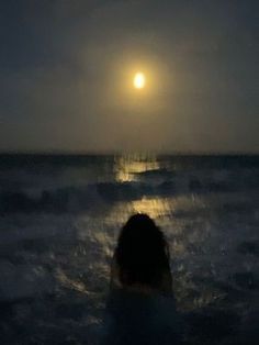 a person standing in the ocean at night looking out into the water with the moon shining behind them