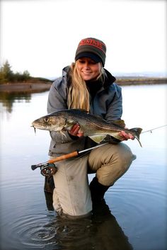 a woman kneeling down in the water holding a fish and fishing rod while wearing a beanie