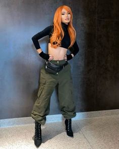 a woman with long red hair is standing in front of a wall wearing green pants and black boots