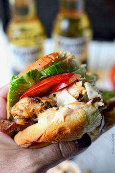 a hand holding a sandwich with chicken, lettuce and tomatoes on it in front of bottles