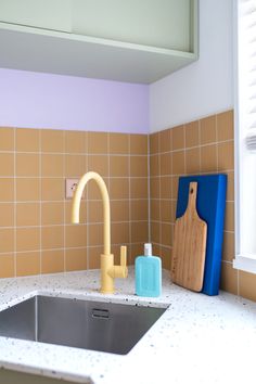 a kitchen sink and cutting board in front of a window with yellow tiles on the wall