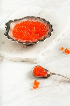 a spoon full of red cavia on a white marble countertop next to a bowl with orange cavia