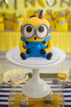 a minion birthday cake sitting on top of a white cake plate with blue and yellow decorations