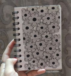 a hand holding a spiral notebook with black dots on the front and back cover, in front of a patterned background