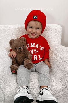 a little boy sitting on top of a white chair holding a teddy bear wearing a red hat